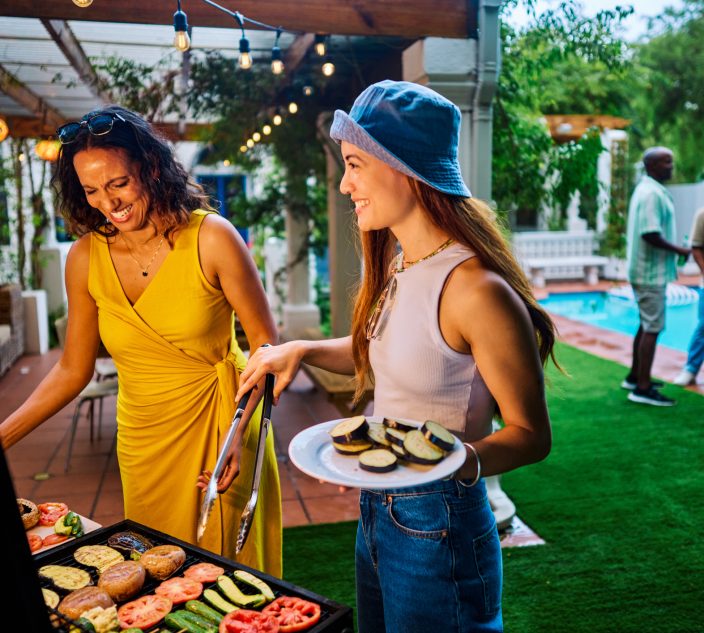 Two women preparing and grilling vegetables at an outdoor barbecue party. Social gathering with food. Summer cooking and celebration concept. Meat free shot.
