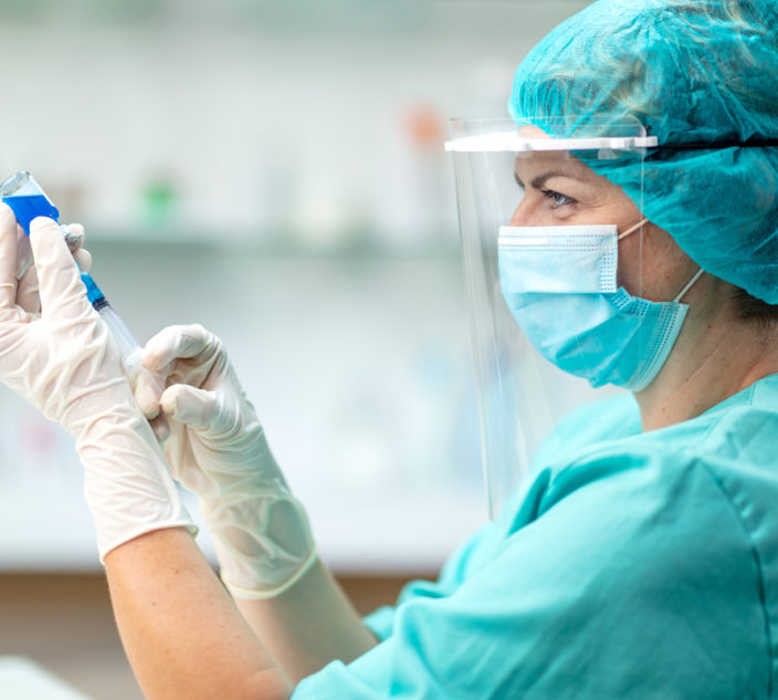 Female doctor pulling COVID-19 vaccine liquid solution from vial with syringe, wearing protective workwear, gloves, face mask and visor.