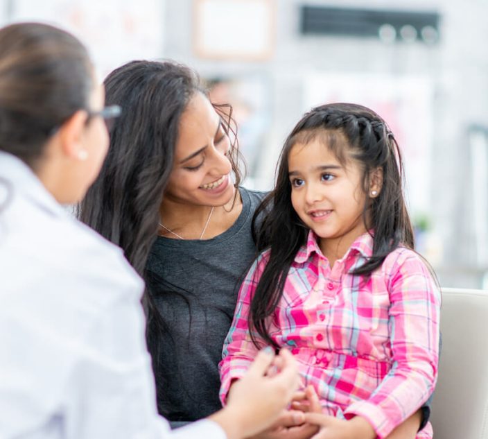 A Mother and her Daughter sit in a medical office across from their Pediatrician during an appointment. The little girl is sitting on her Mothers lap and is smiling as they talk with the doctor.