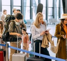 Passengers stand in a busy airport terminal, waiting in line for check-in and boarding while carrying luggage.