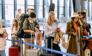 Passengers stand in a busy airport terminal, waiting in line for check-in and boarding while carrying luggage.